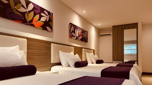 Habitación de hotel con tres camas, cada una con ropa blanca y detalles morados. Se observan obras de arte abstractas con motivos florales sobre las camas.