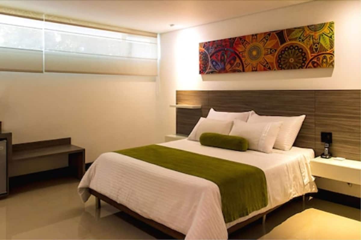 A modern hotel room with a large bed, a nightstand, a window with blinds, and a colorful abstract painting on the wall.