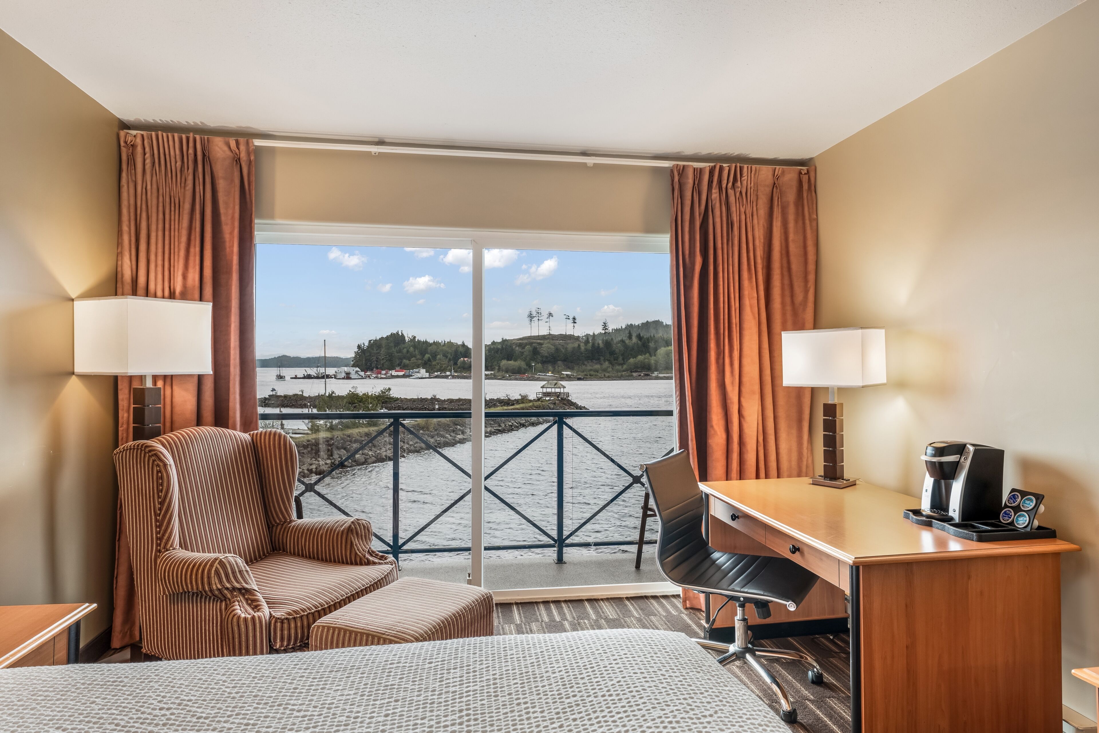 A hotel room with a bed, two chairs, a desk, a coffee maker, and a view of a body of water.