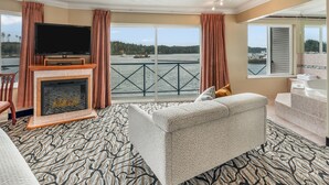A living room with a fireplace, a flat-screen TV, a sofa, and a view of the water. - Glen Lyon Inn & Suites (Port Hardy)