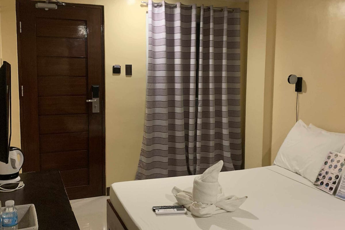 Ein Hotelzimmer mit Holztür, einem Bett mit weißen Laken, einer Fernbedienung, einer Wasserflasche und einem Handtuch auf dem Bett.