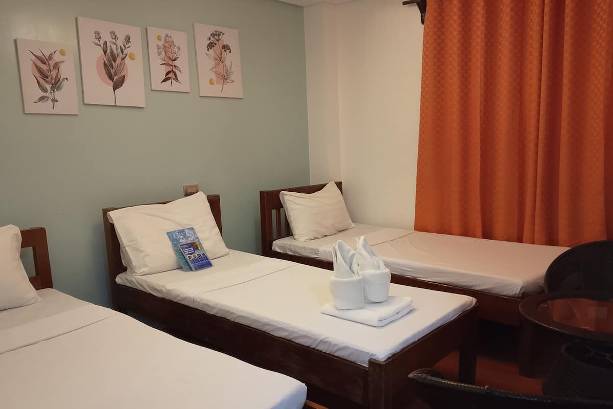 Ein Hotelzimmer mit drei Einzelbetten, jedes mit weißen Laken und Kissen. Es gibt einen kleinen Tisch mit einem Stuhl, ein blaues Informationsheft und einen Korb für Handtücher. An den Wänden hängen drei botanische Drucke.