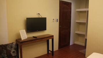 Quarto com televisão na parede, um banco de madeira, uma porta e uma estante branca.