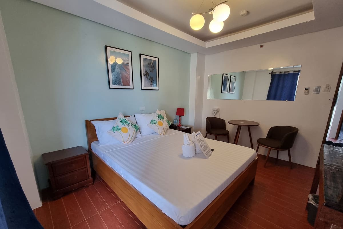 Ein helles Hotelzimmer mit einem großen Bett, einem kleinen Schreibtisch, einem Sessel und einem runden Tisch. An der Wand befinden sich zwei gerahmte Bilder, ein Spiegel und eine rote Lampe auf dem Schreibtisch.