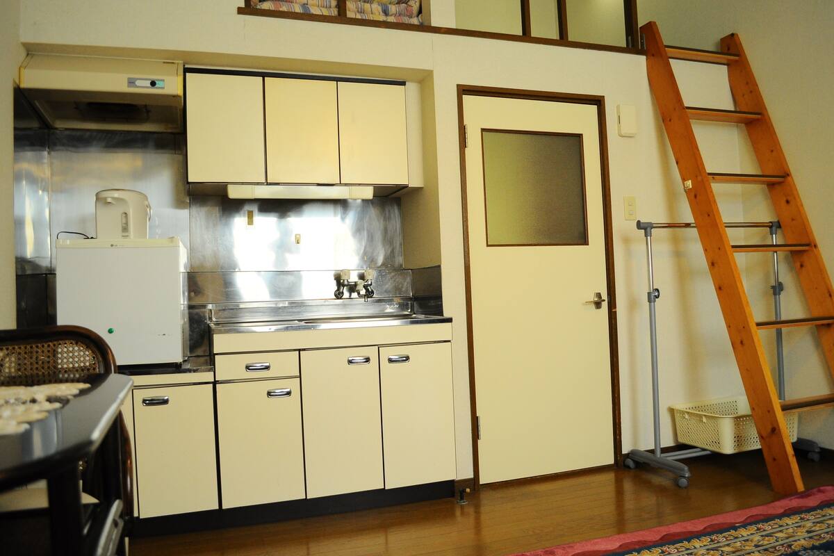 A compact kitchen with a refrigerator, stove, and cabinets. A wooden ladder is leaning against the wall.