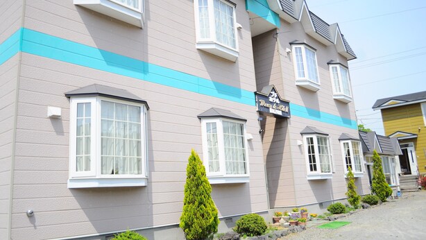 A multi-story hotel with a light-colored facade and blue trim. It features several windows, a sign that reads "Hotel to School," and a small garden area with plants and a pathway. - Petit Hotel Koizumi (Toyako)