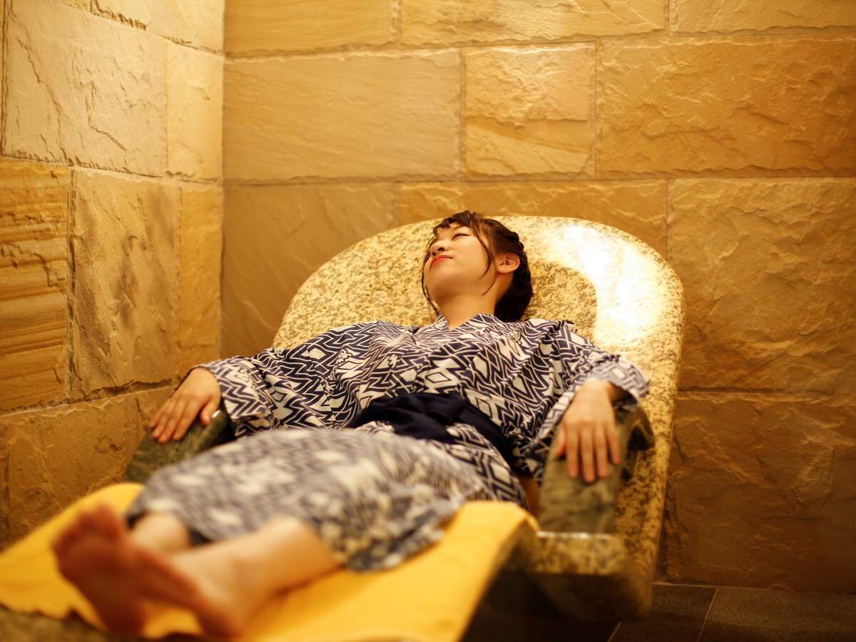 A person lying in a chair with a patterned blanket, in a room with stone walls.