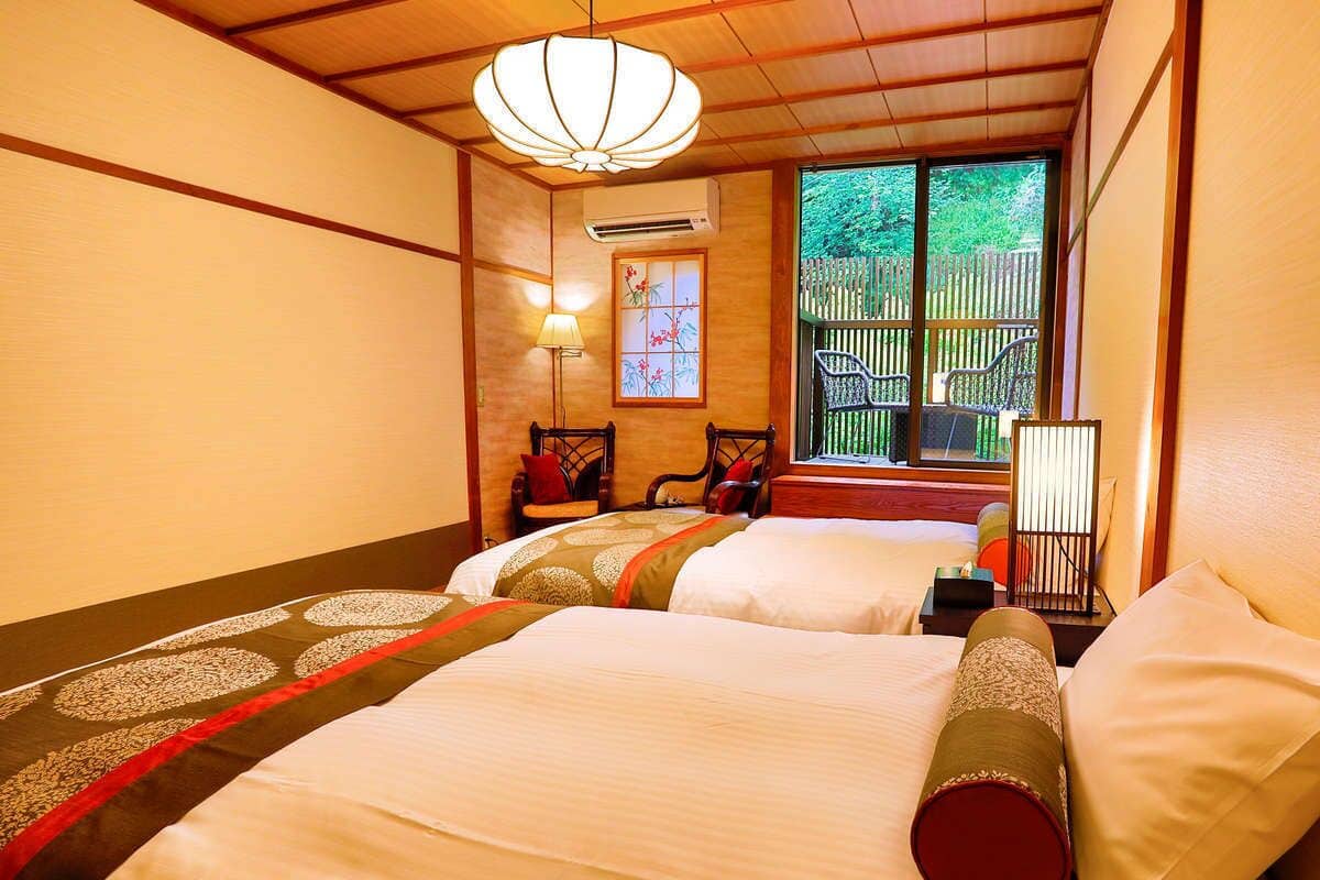 A hotel room with two beds, a wooden ceiling, a window with a view of greenery, a chair, and a lamp.