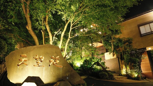 Un monumento in pietra con caratteri giapponesi, circondato da alberi e un edificio con persiane di legno.