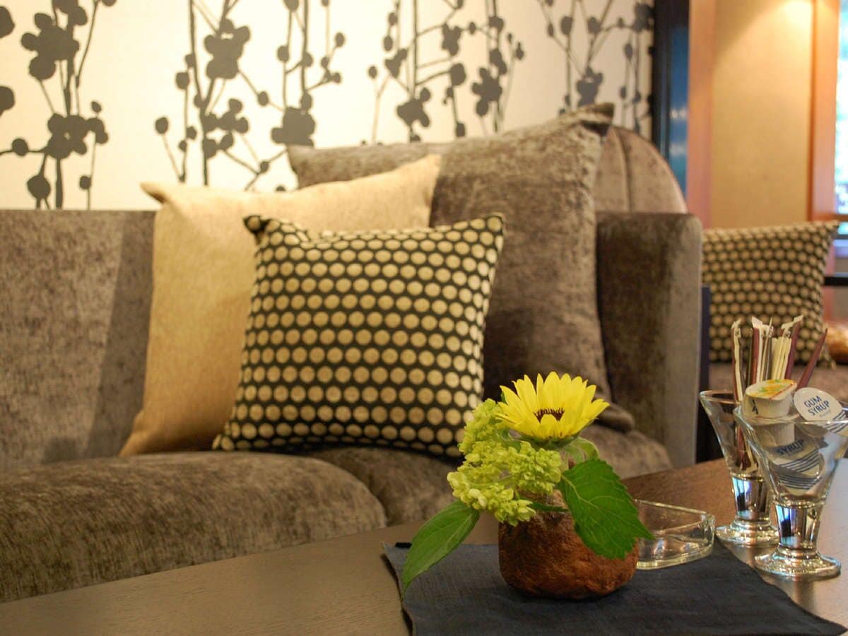 A living room with a sofa, patterned pillows, a small table with a vase of flowers, and a glass of water.