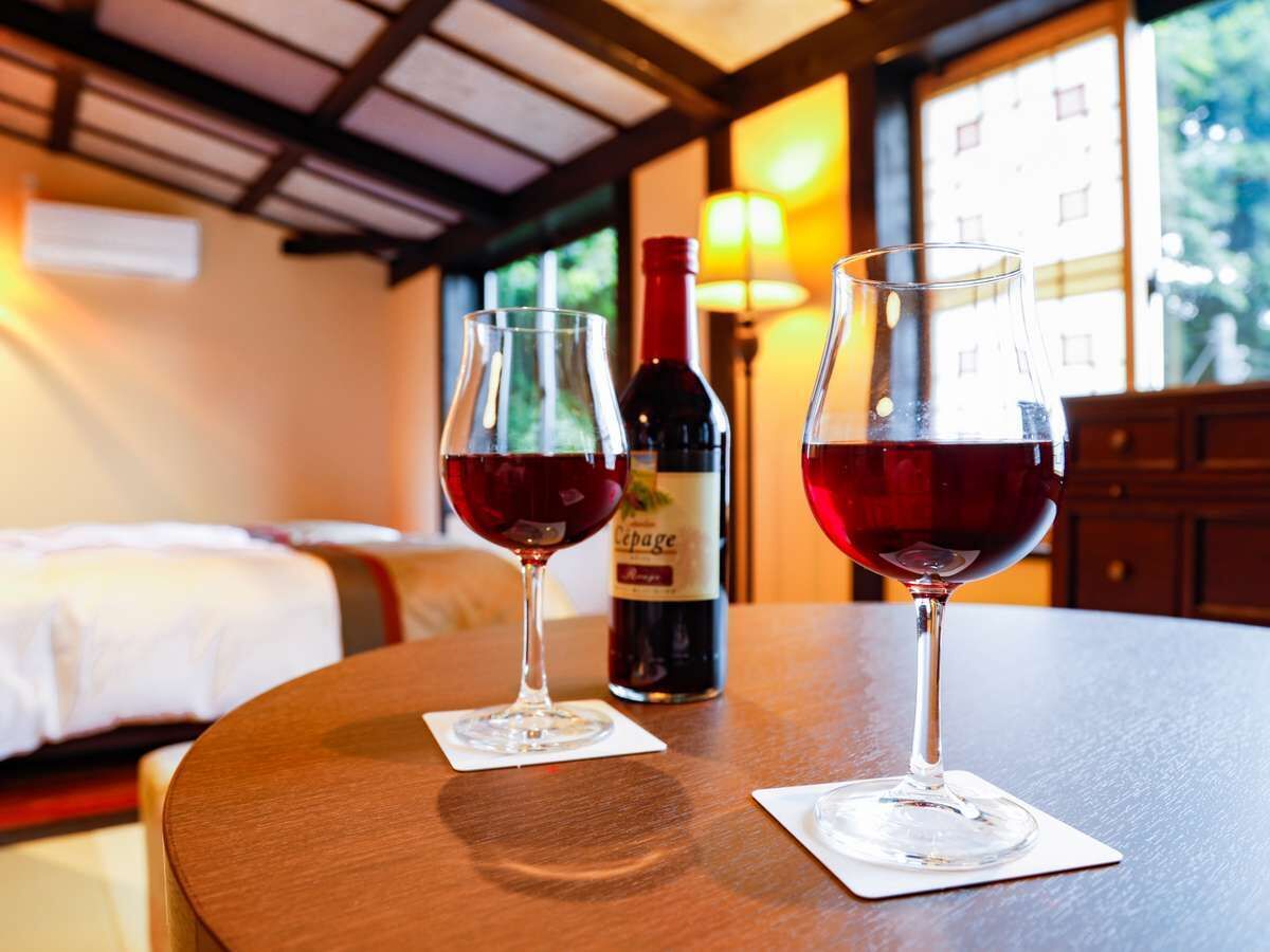 A wooden table with two wine glasses and a bottle of wine in a room with a bed and a lamp in the background.