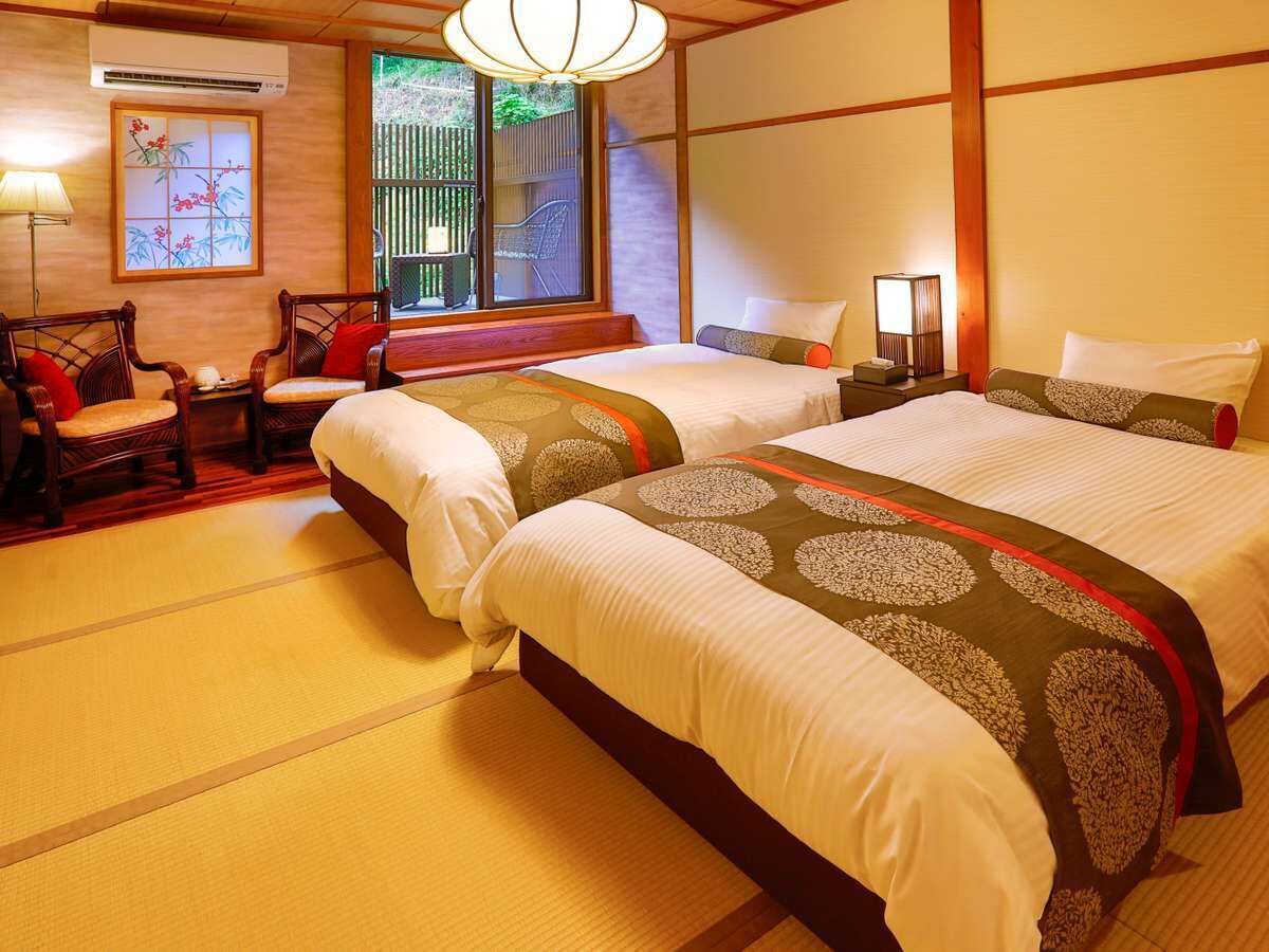 Habitación de hotel tradicional japonesa, con suelo de tatami, dos camas con ropa de cama estampada, cabecero de madera, ventana con vista a zonas verdes y una pequeña mesa con sillas.