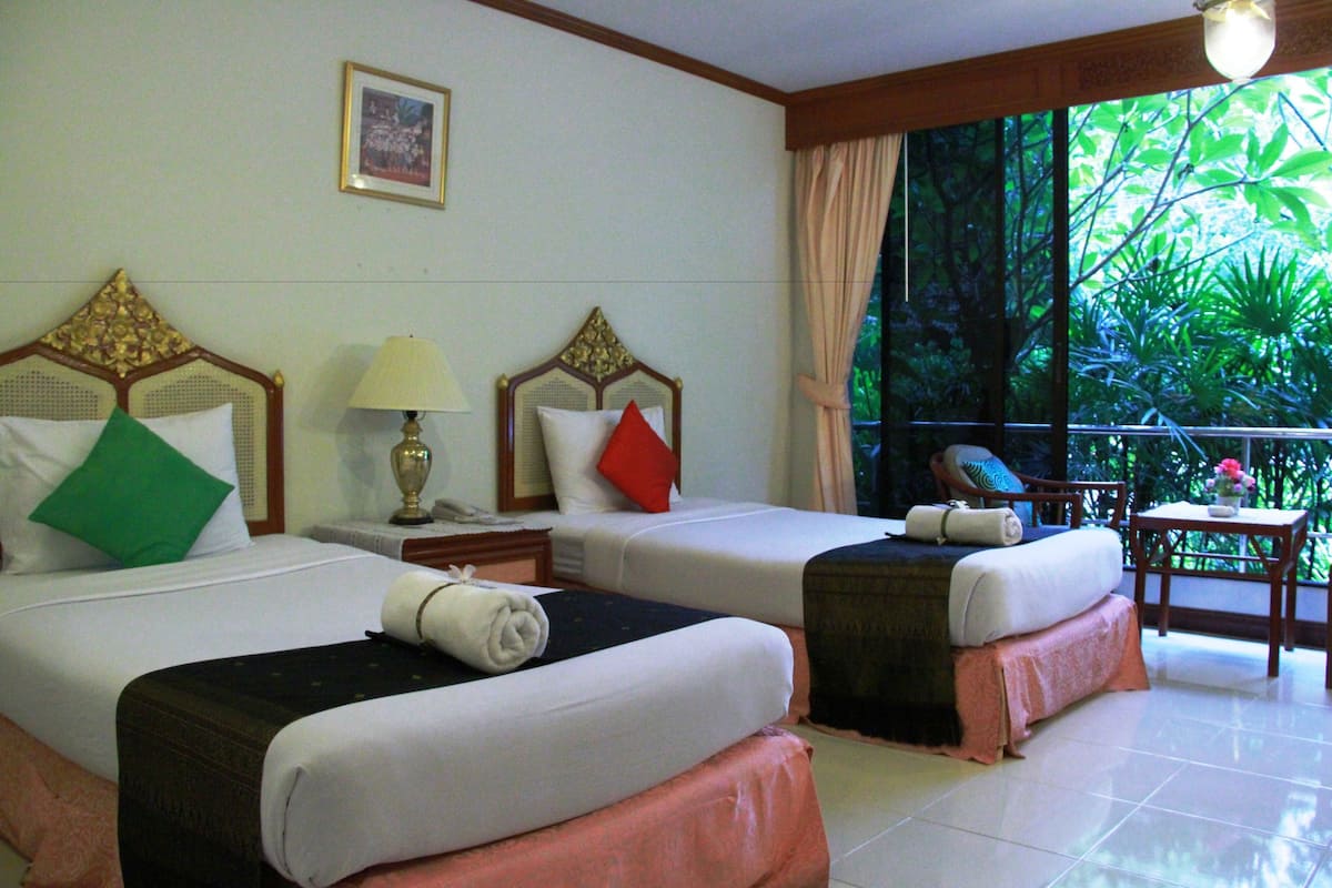 A hotel room with two beds, a nightstand, a lamp, and a view of greenery through the window.