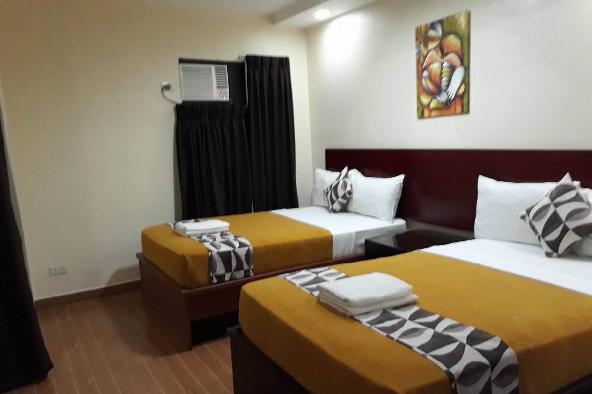 Une chambre d’hôtel avec deux lits, une grande fenêtre avec des rideaux noirs, un appareil de climatisation et un tableau coloré accroché au mur.