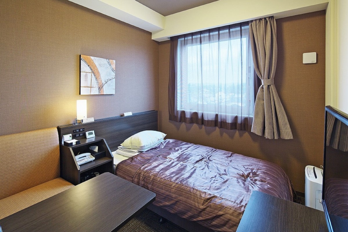 Habitación de hotel con cama, escritorio, silla, mesita de noche y ventana con cortinas.