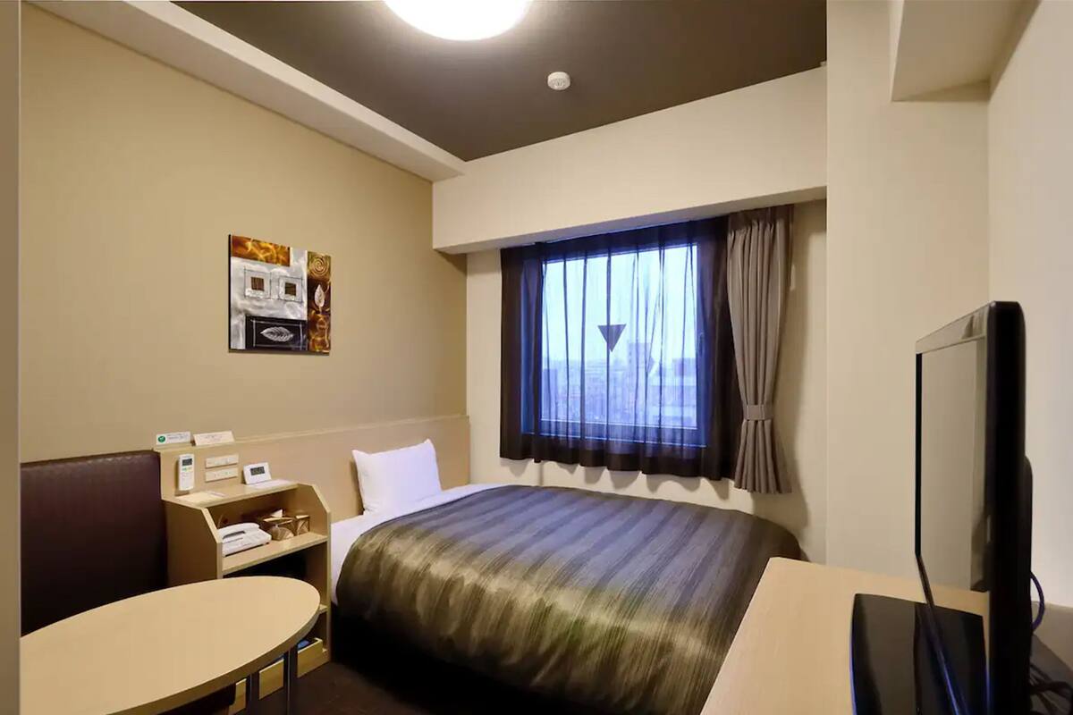 Una habitación de hotel con cama, escritorio, silla, televisor y ventana con cortinas.