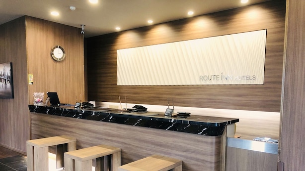 Un moderno sector de recepción de hotel con mostrador de mármol, taburetes de madera, un reloj y un cartel que dice "ROUTE IN HOTELS".