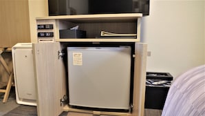 Un refrigerador compacto en una habitación de hotel, un cesto de basura pequeño y una cama con manta a rayas.
