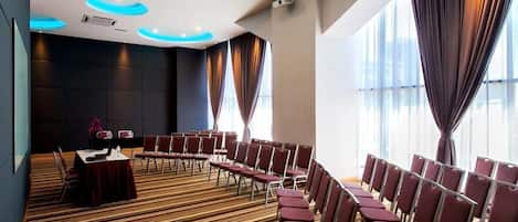Una sala conferenze con tappeto a righe, file di sedie, un tavolo con tovaglia rossa e ampie finestre con tende.