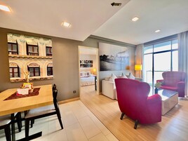 A modern hotel room with a dining area, a living room with a red armchair, a sofa, and a large window offering a view of the outside.