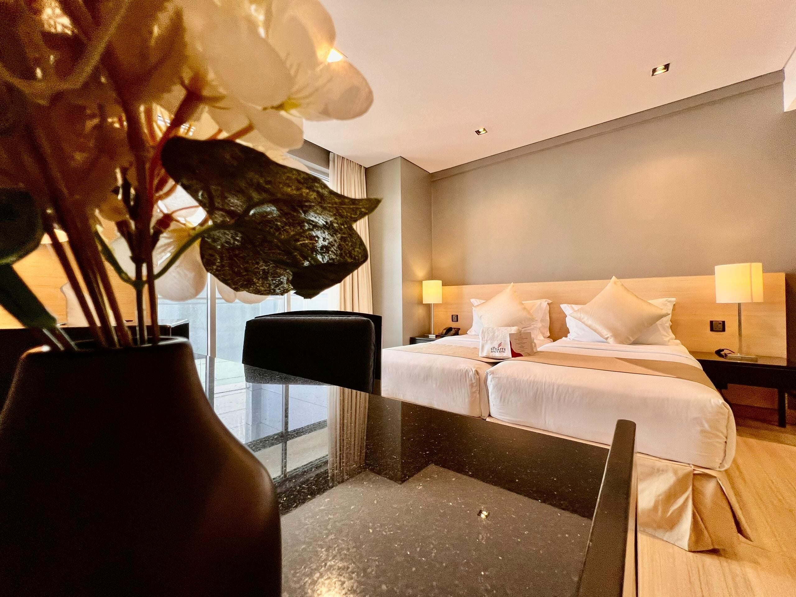 Quarto de hotel com cama, luminárias de cabeceira, um vaso com flores e vista para o exterior.