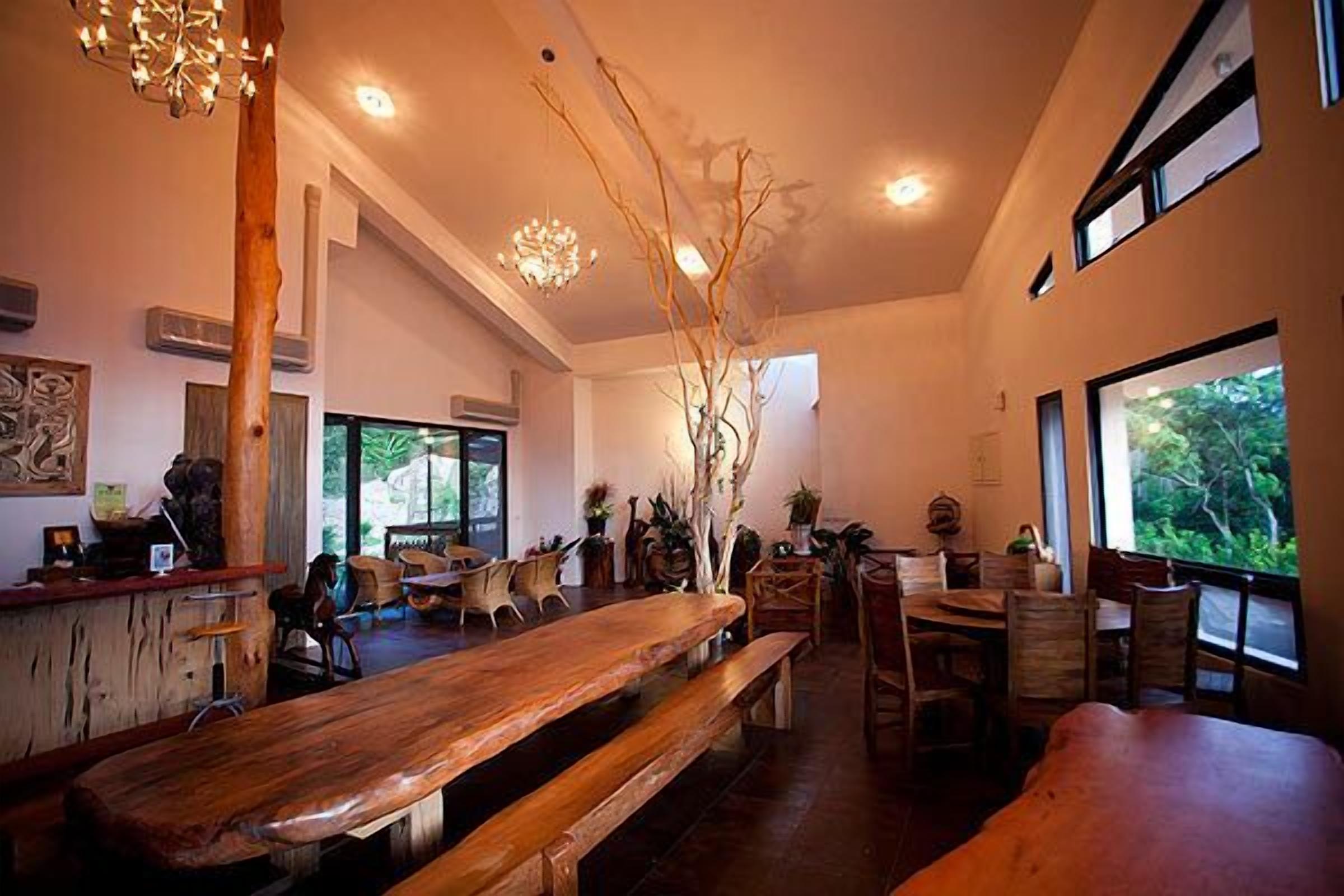 Un restaurante amplio con bancos y mesas de madera, ventanas grandes que ofrecen vistas a zonas verdes, y un candelabro colgando del techo.