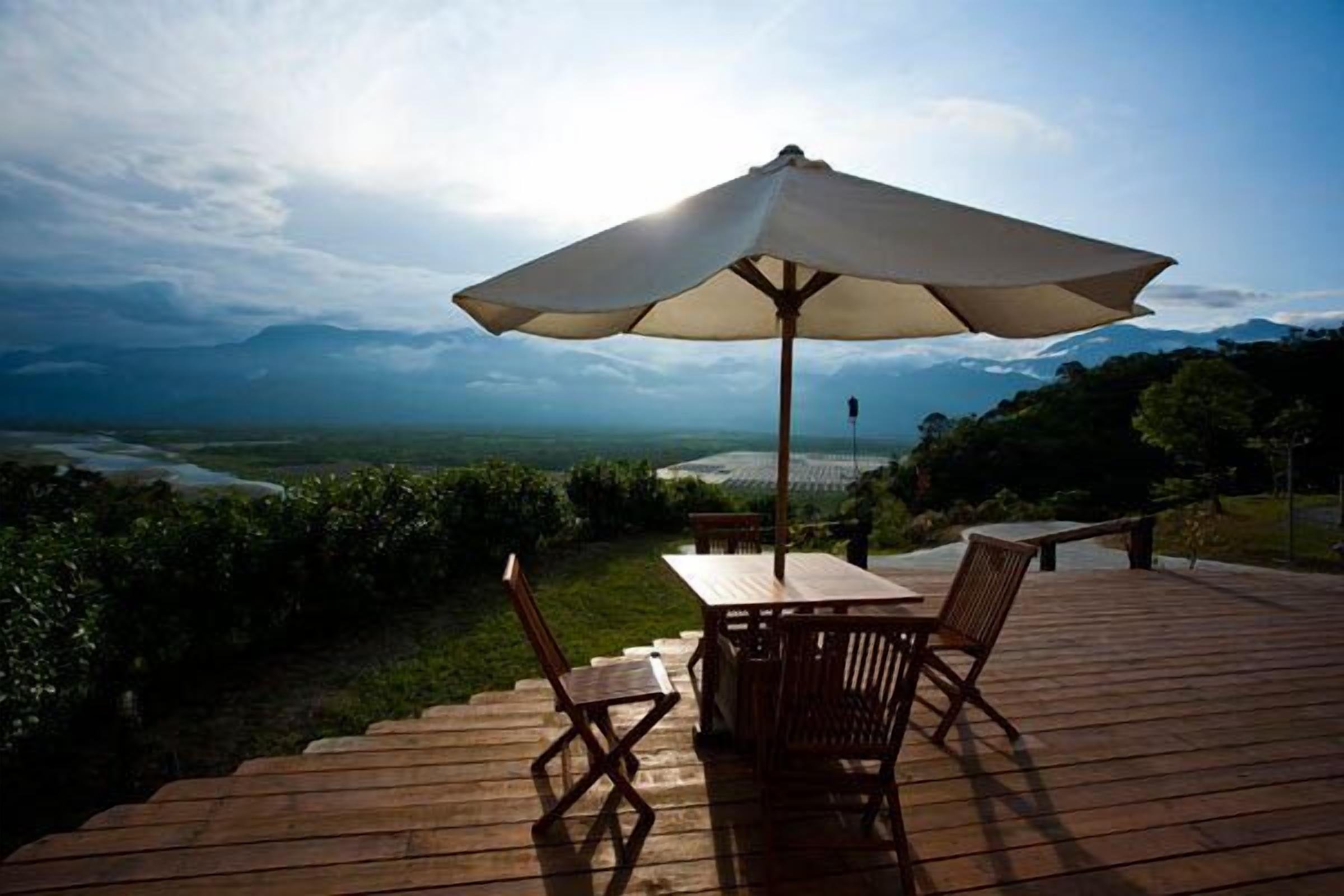 Una terraza de madera con una mesa y sillas bajo un paraguas grande, con vistas a un paisaje pintoresco con montañas y un río.