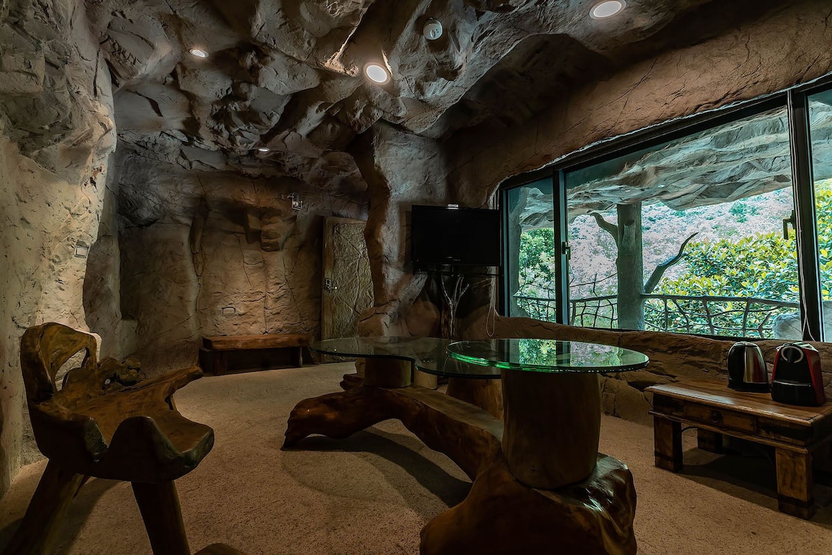 A room with a natural stone ceiling, a glass table, wooden chairs, and a large window with a view of greenery.