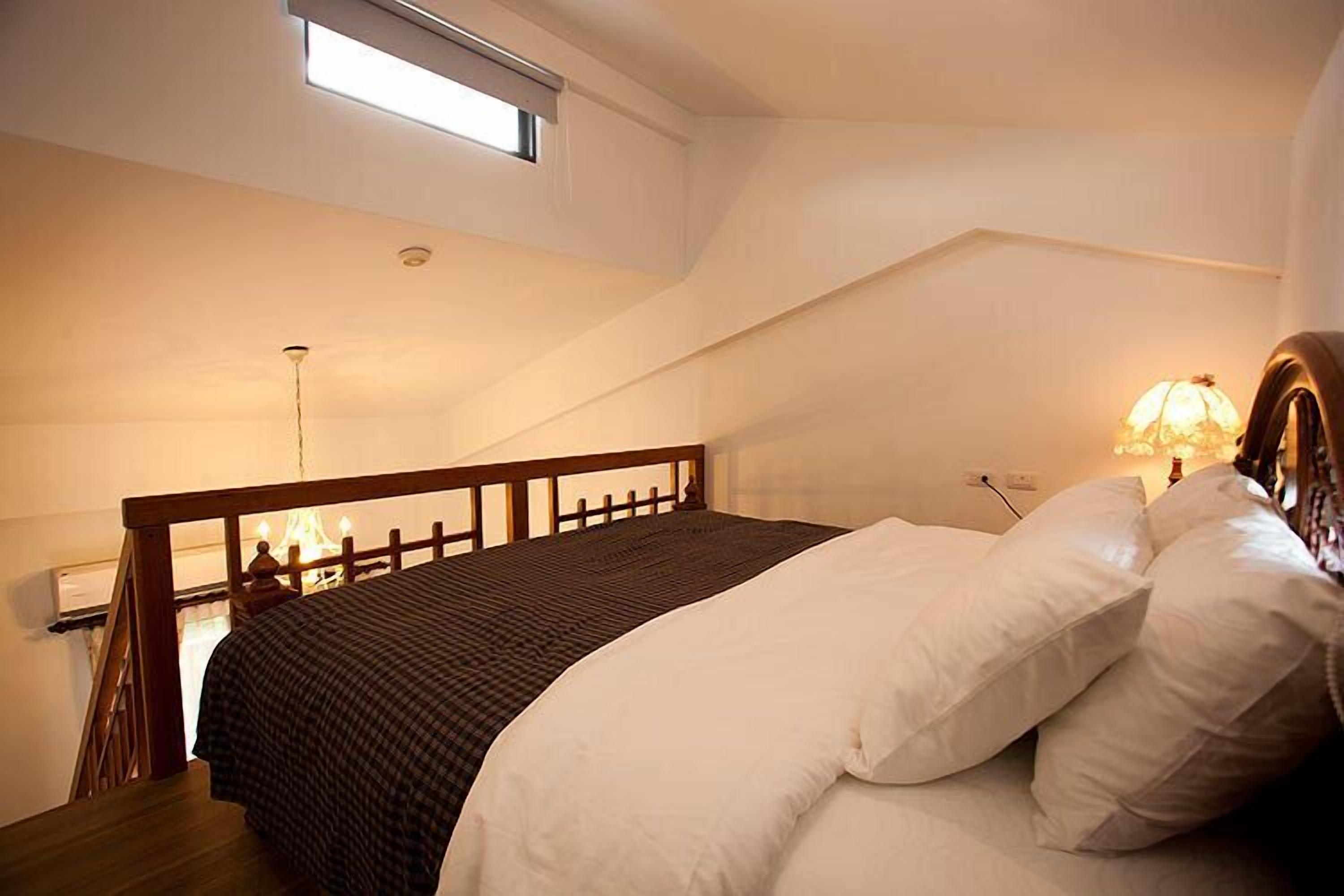 Un dormitorio con estructura de cama de madera, colcha a cuadros y almohadas blancas. Hay una ventana sobre la cama y una lámpara en la mesita de noche.