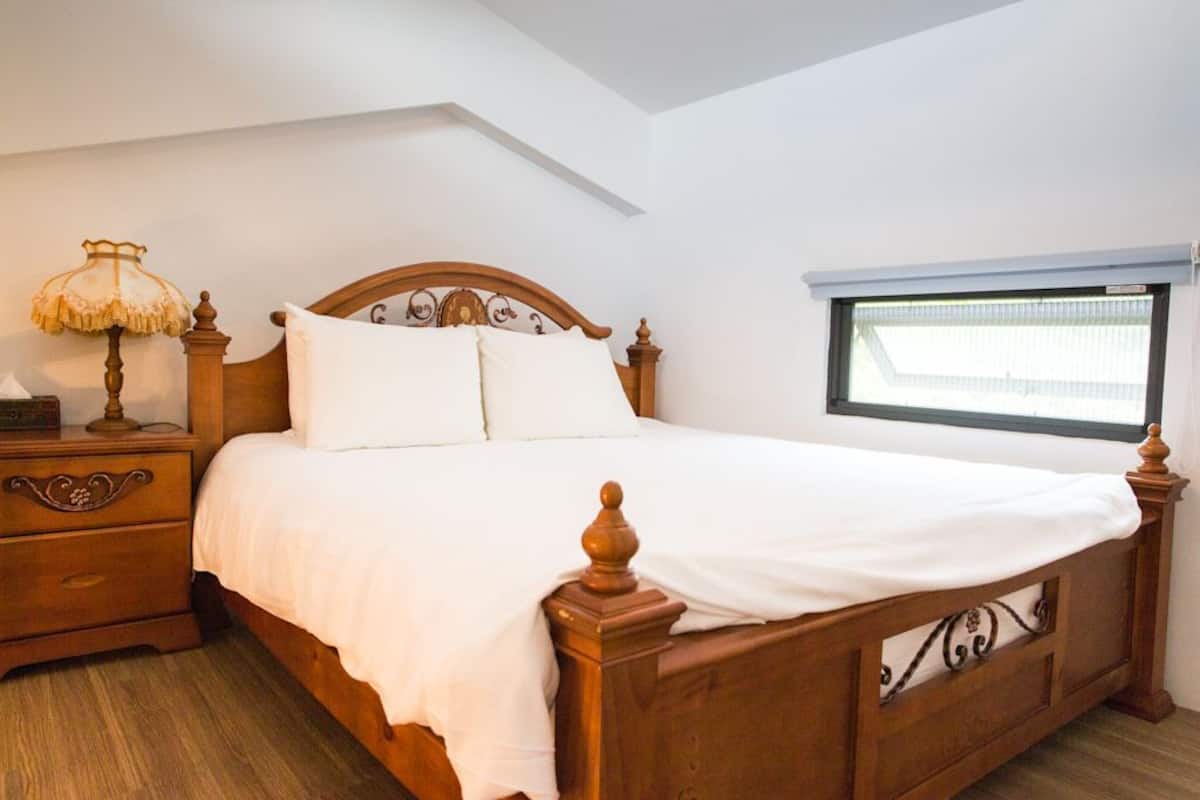 A wooden bed with white bedding in a room with a sloped ceiling and a window with blinds.