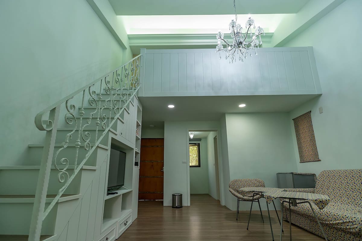 A spacious living area with a high ceiling, a staircase with ornate railings, a chandelier, and a television.