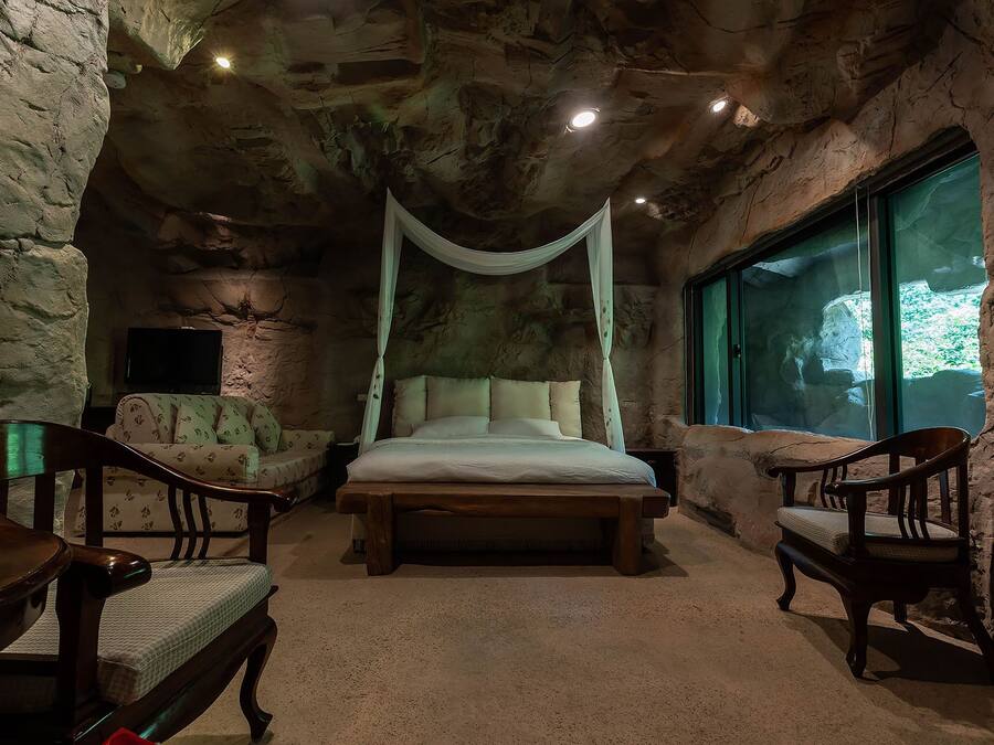 A stone-walled bedroom with a canopy bed, two wooden chairs, and a large window.