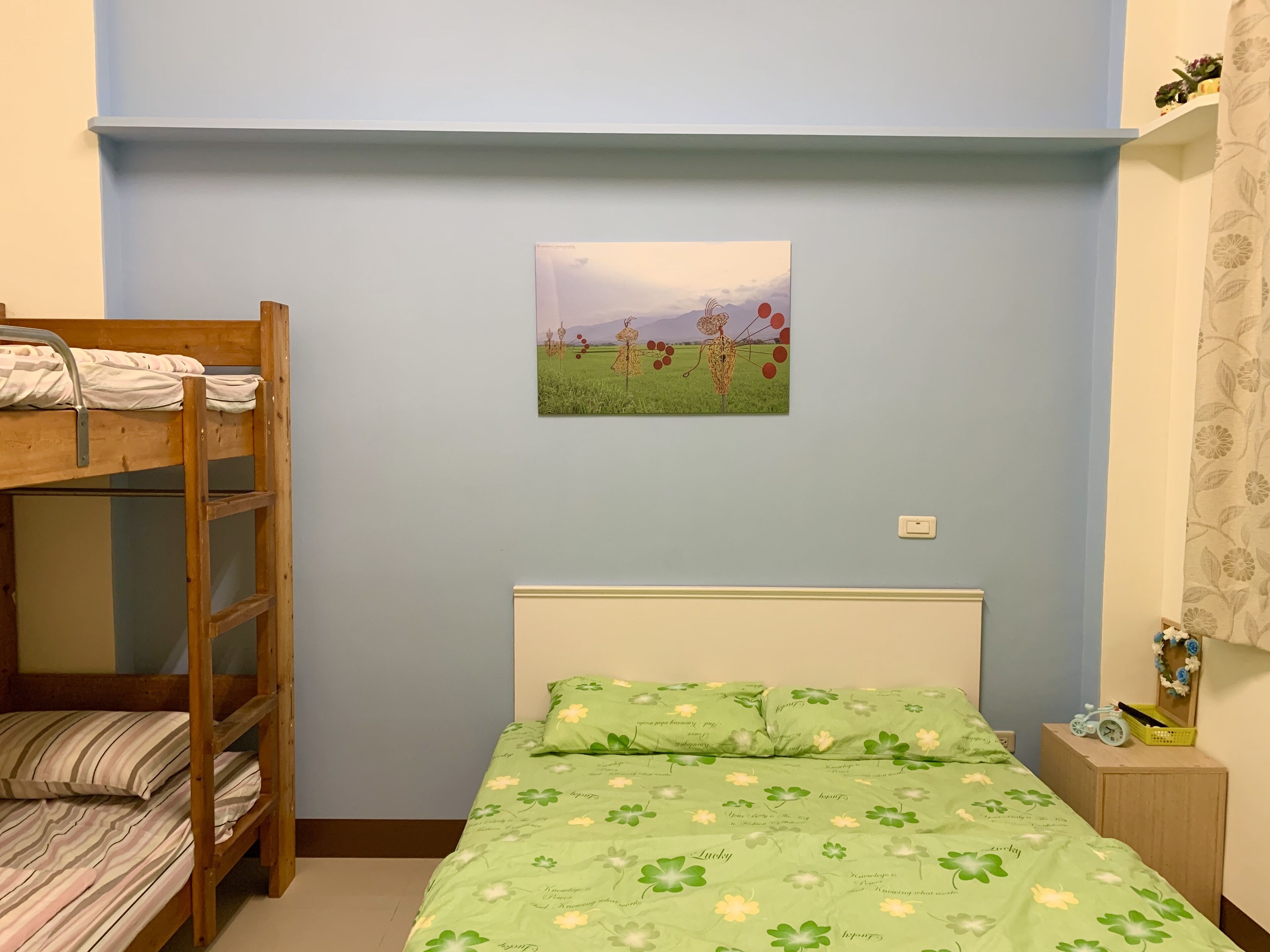 Una habitación con una cama litera, una cama individual con colcha floral verde, un cuadro colgado en la pared de un campo con globos rojos, y una mesita de noche pequeña con un reloj y una cesta.