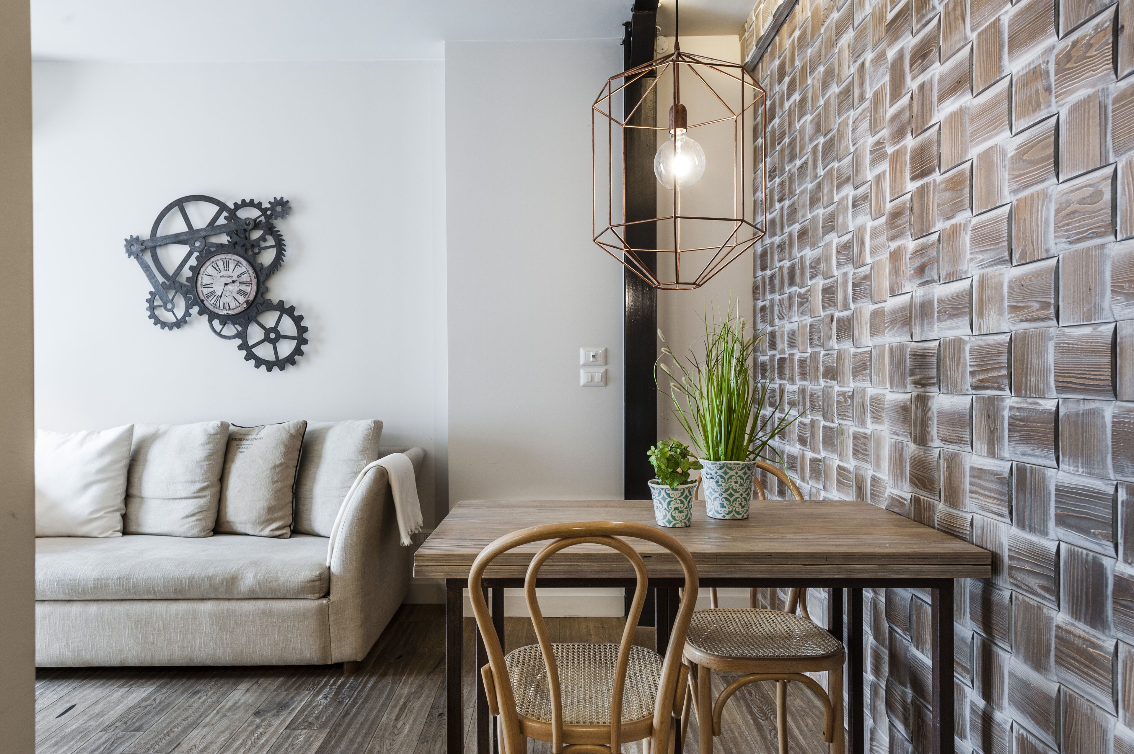 A dining area with a wooden table, two wicker chairs, a wall clock, a hanging light fixture, and a textured wall.