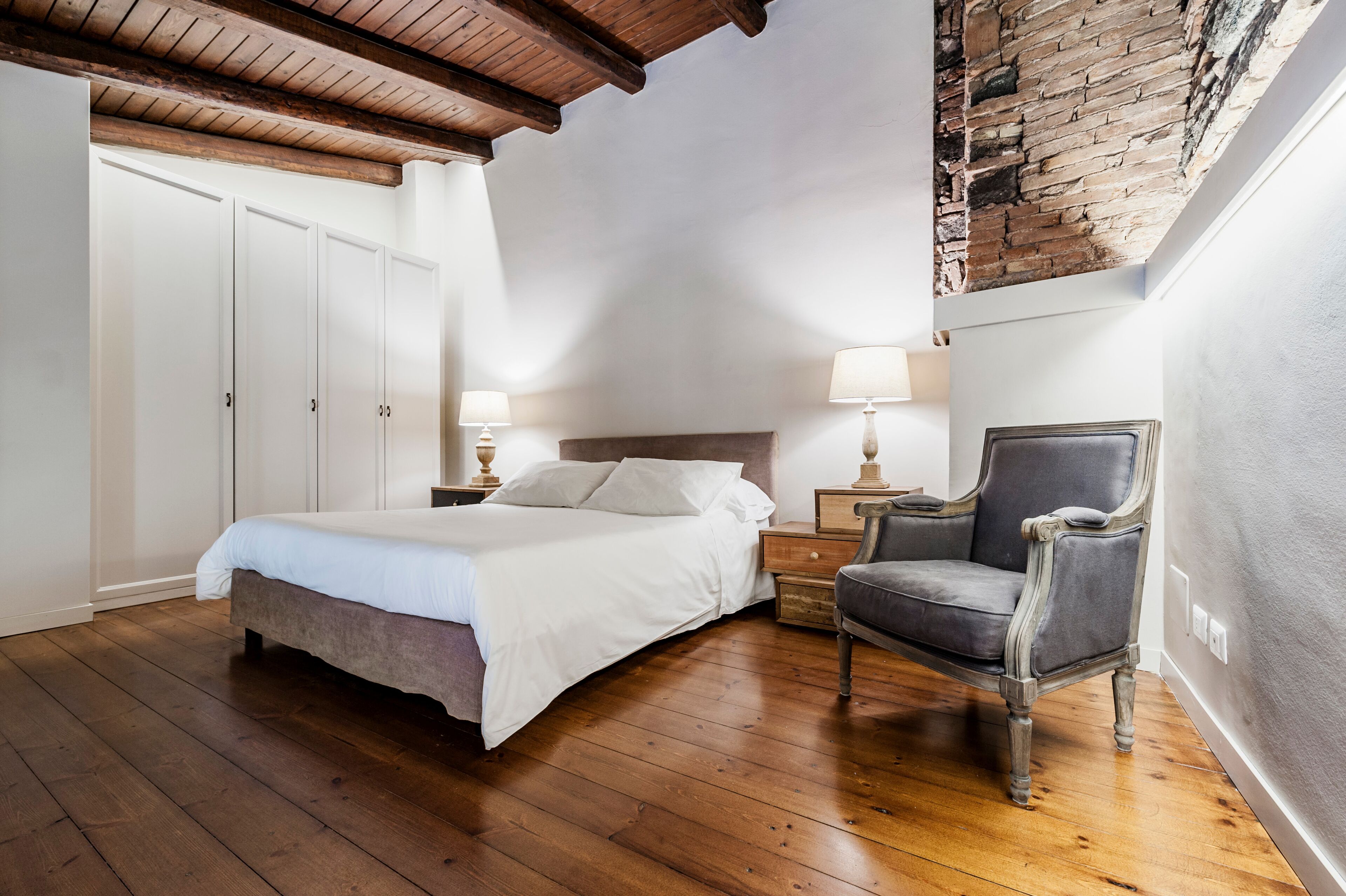 A bedroom with a wooden floor, a bed, a chair, and a nightstand with lamps.