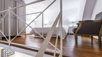 A bedroom with a wooden floor, a bed with white bedding, a chair, and a nightstand with a lamp.