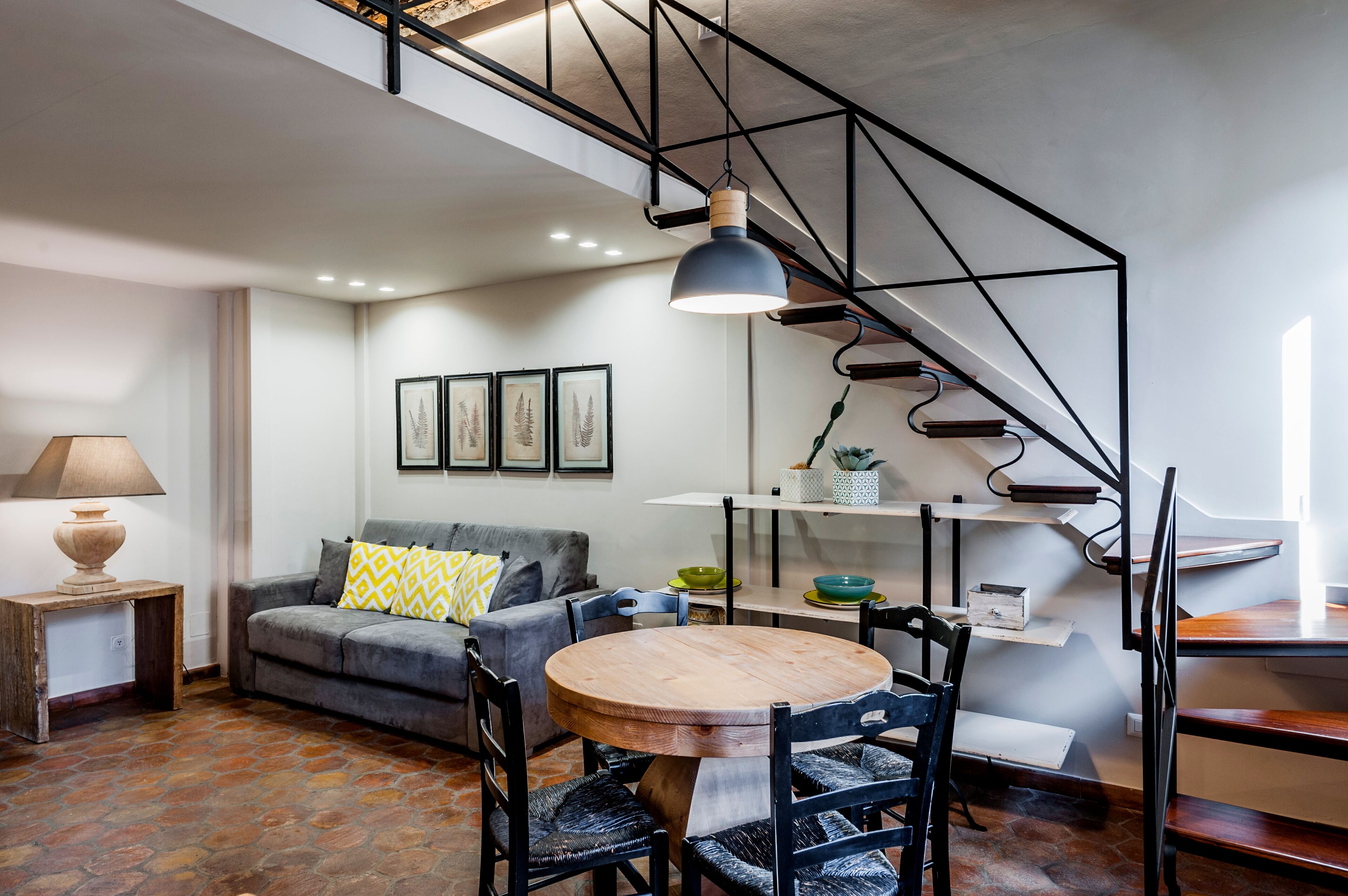 A modern living room with a staircase, a round wooden table, and a grey sofa with patterned cushions.
