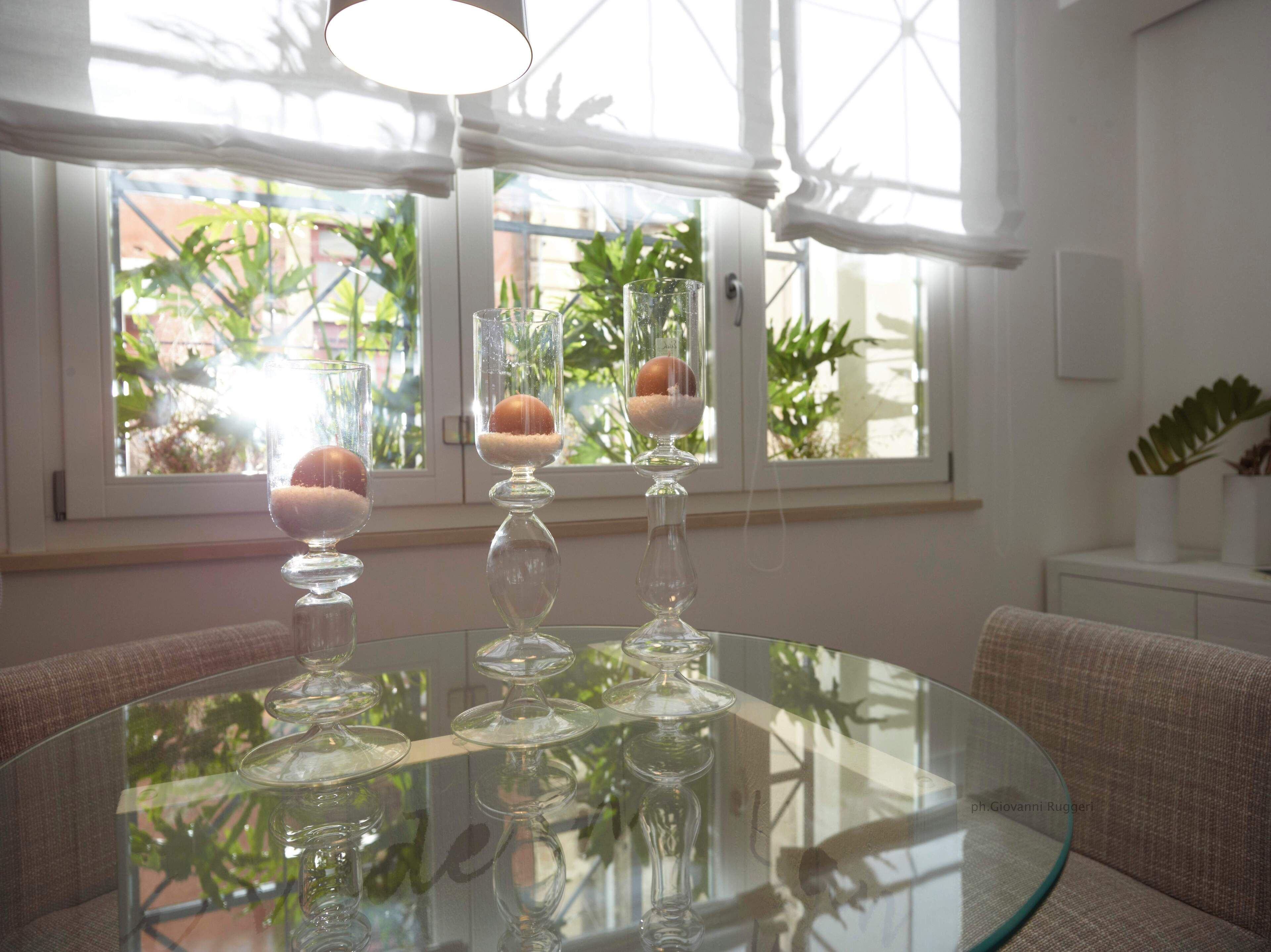 Ein runder Glastisch mit drei klaren Glassäulen, in denen jeweils eine kugelförmige Form steckt. Der Raum verfügt über ein Fenster mit weißen Vorhängen, eine Couch und eine kleine, weiße Kommode mit einer Pflanze.