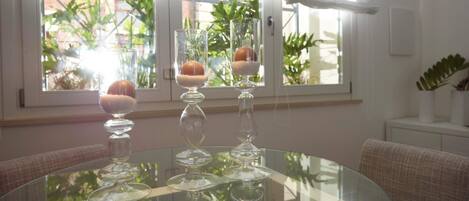 Ein runder Glastisch mit drei klaren Glassäulen, in denen jeweils eine kugelförmige Form steckt. Der Raum verfügt über ein Fenster mit weißen Vorhängen, eine Couch und eine kleine, weiße Kommode mit einer Pflanze.