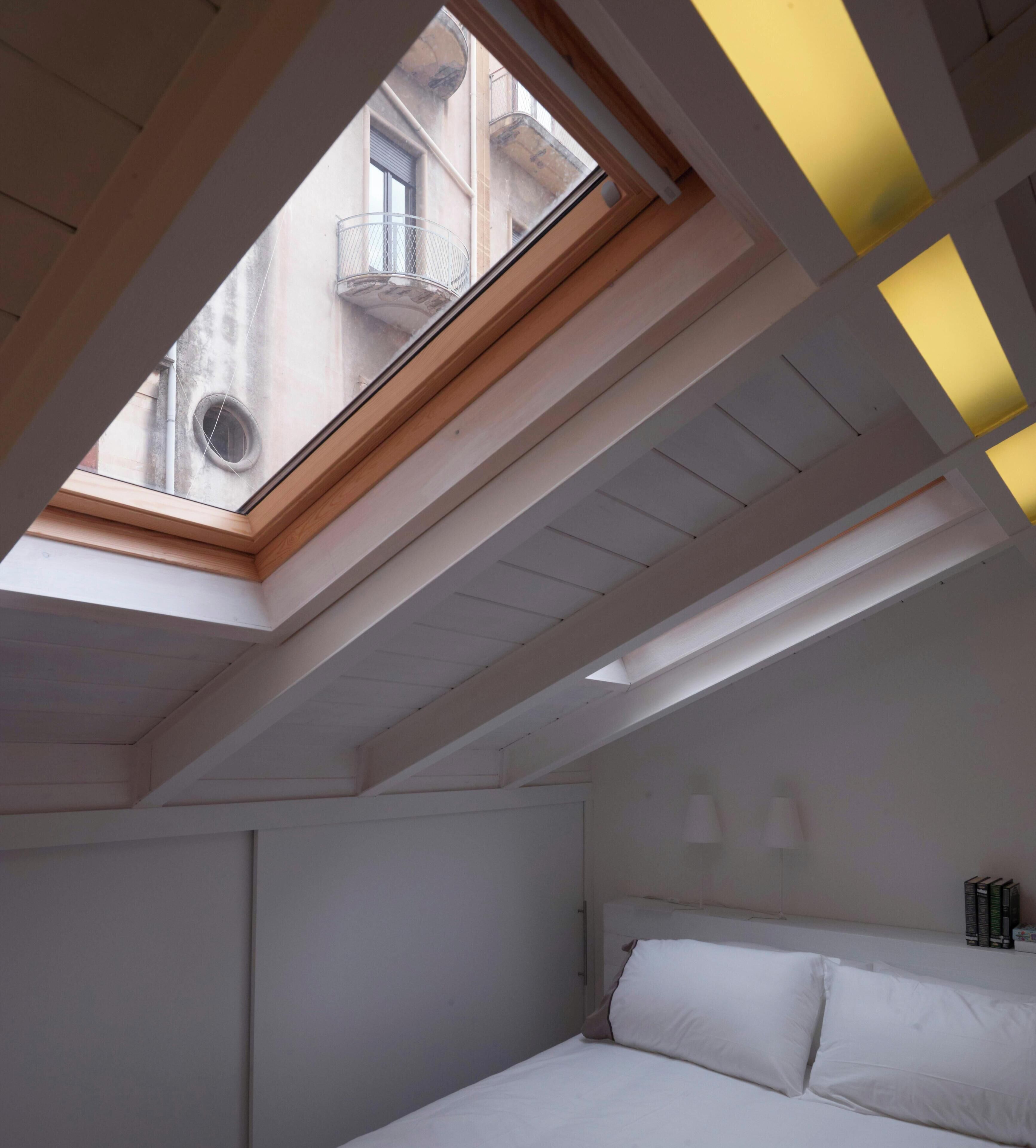 A bedroom with a skylight, a bed, and a nightstand with books.