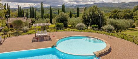 A swimming pool with a sun terrace, surrounded by a fence and trees, overlooking a mountainous landscape.