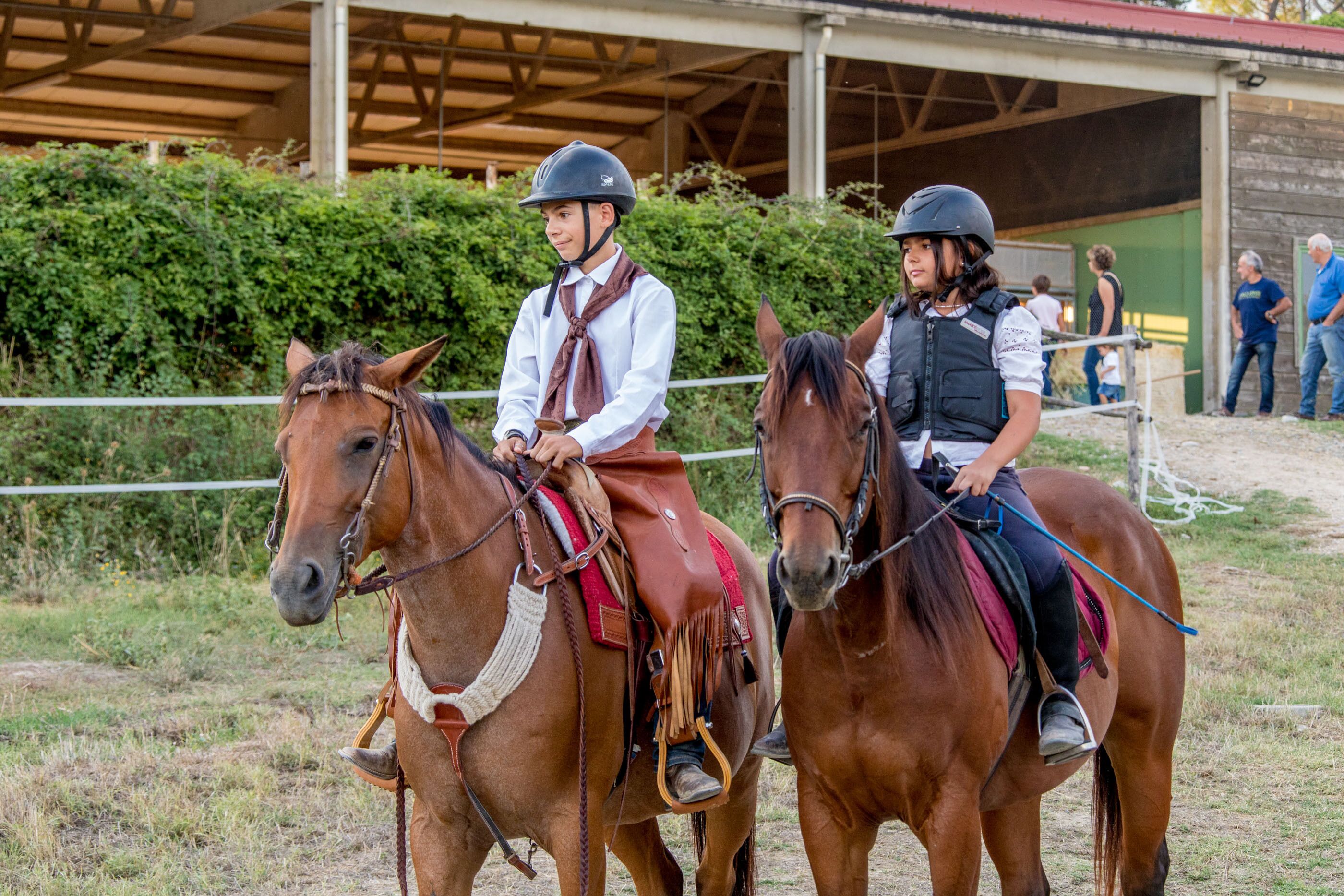 Dos jinetes a caballo en una arena al aire libre, con un edificio techado al fondo.