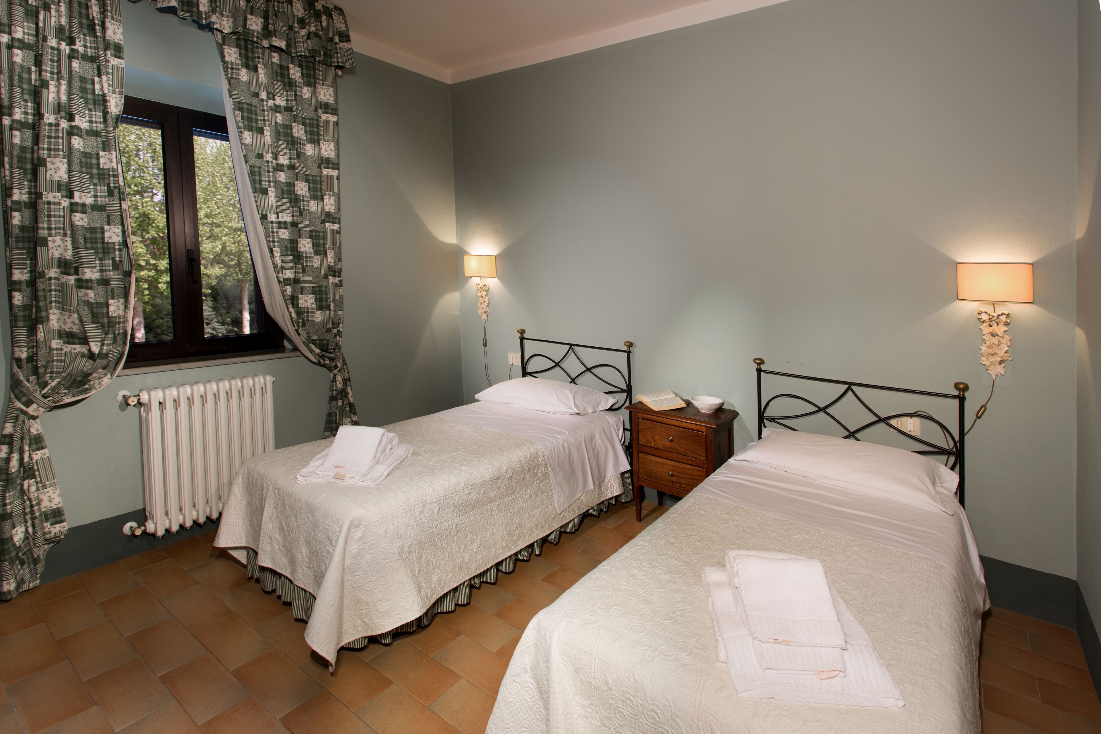 A bedroom with two beds, a window with curtains, a radiator, and bedside lamps.