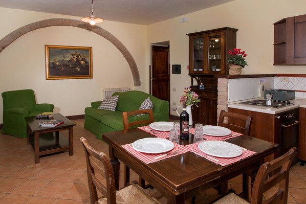 A dining area with a wooden table set for four, a green sofa, a small coffee table, and a kitchen in the background.