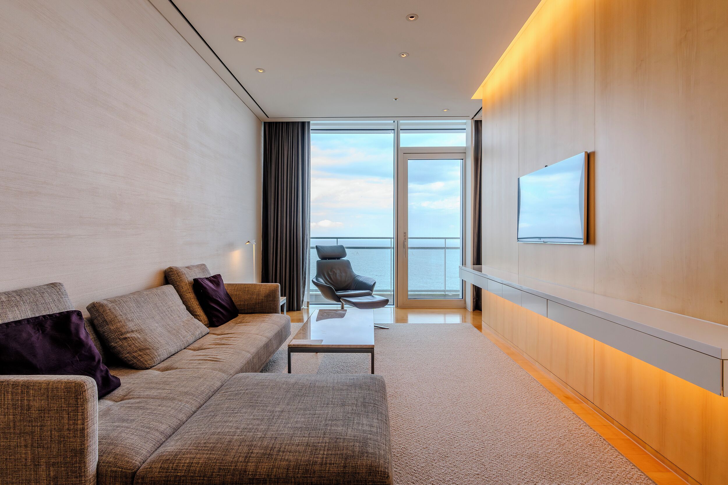 A modern hotel room with a sofa, a coffee table, and a large window offering a view of the sea.