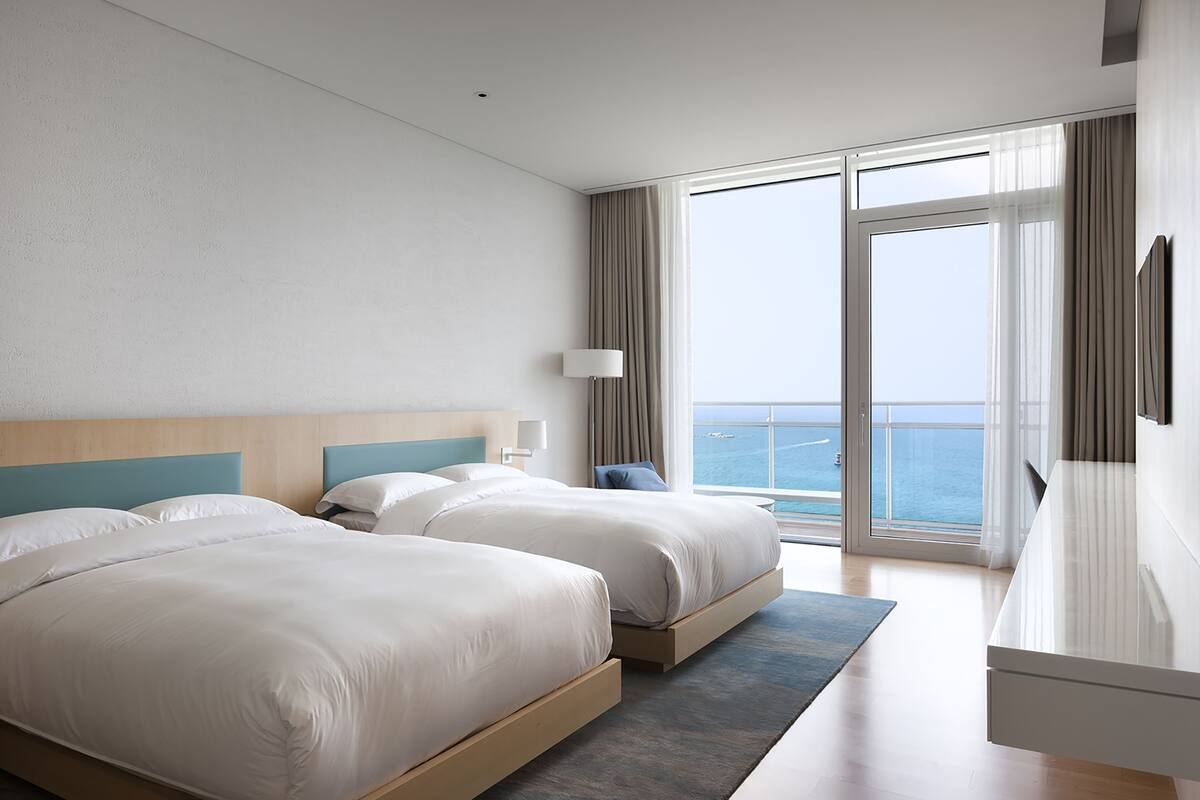Quarto de hotel com duas camas, uma janela grande com cortinas, vista para o mar e uma TV de tela plana na parede.