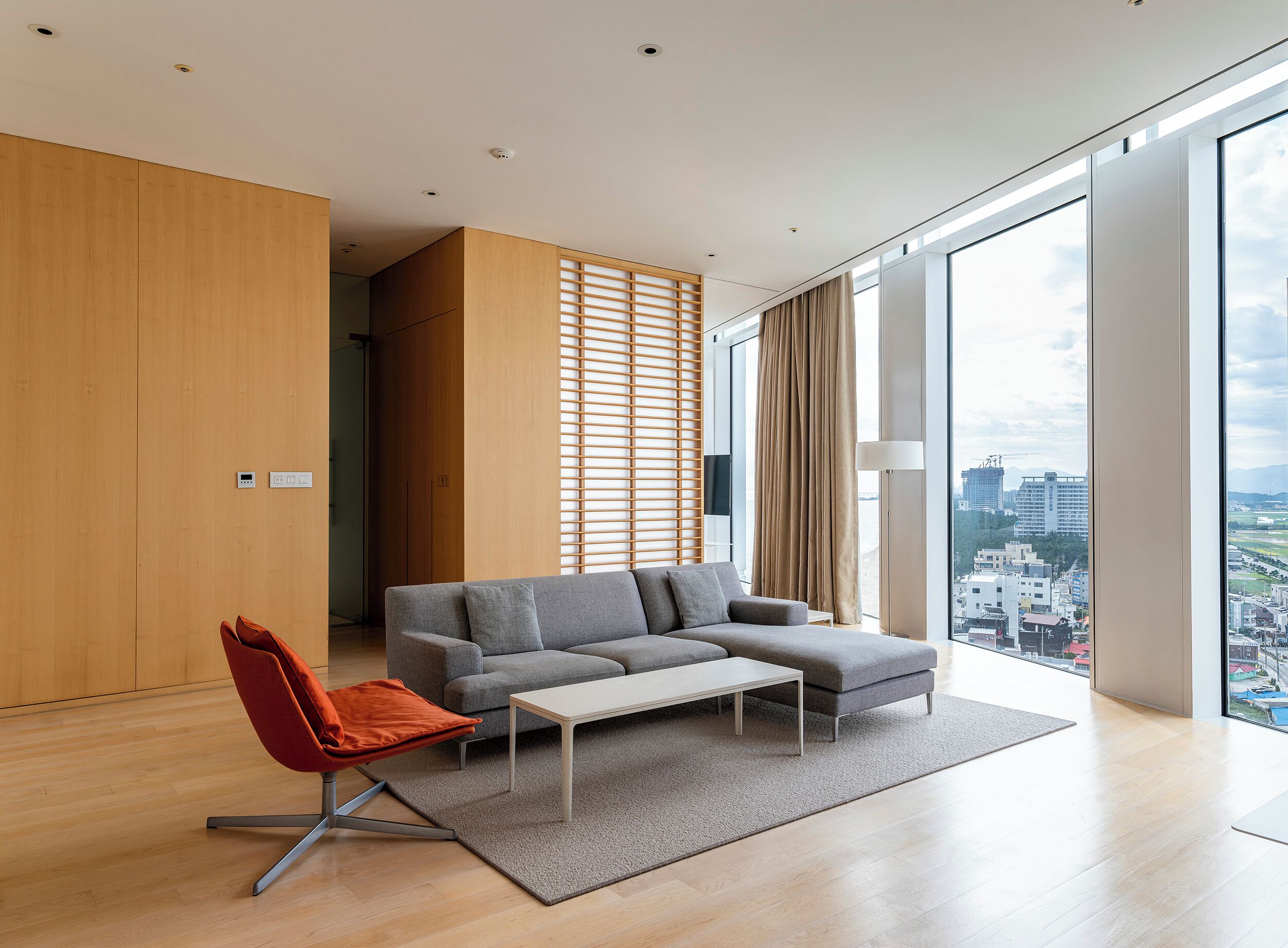 A modern living room with a sectional sofa, a red chair, a coffee table, and a large window offering a city view.