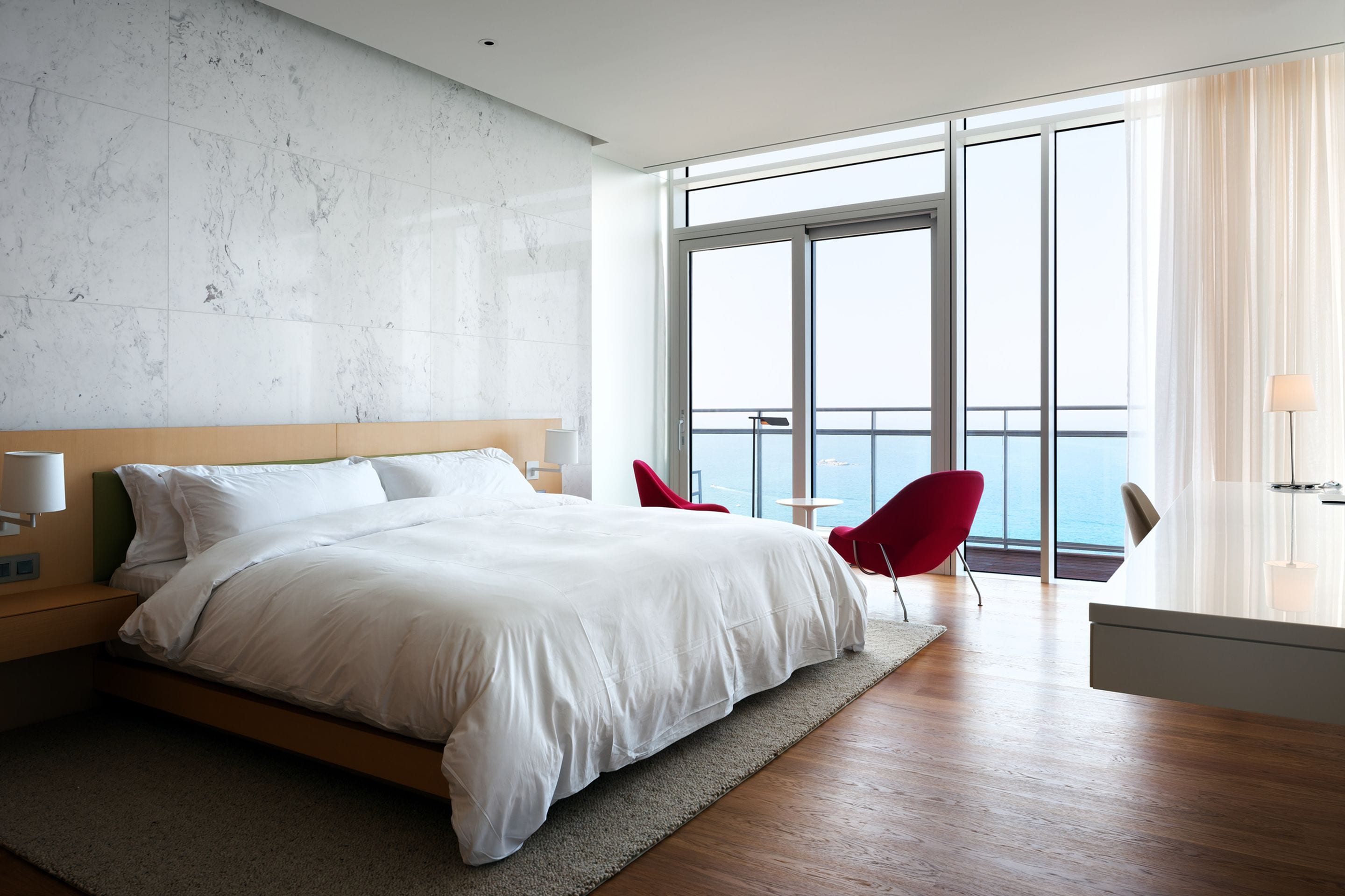 A modern hotel room with a large bed, a view of the ocean, a balcony with red chairs, and a minimalist design.
