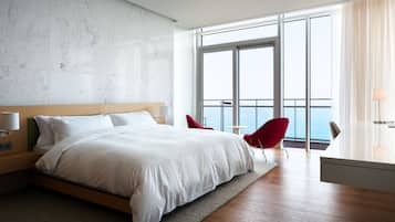 A modern hotel room with a large bed, a view of the ocean, a balcony with red chairs, and a minimalist design.