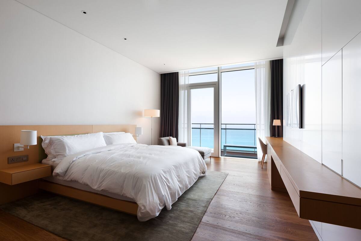 Quarto de hotel moderno com uma cama grande, mesas de cabeceira, escrivaninha e vista para o mar.